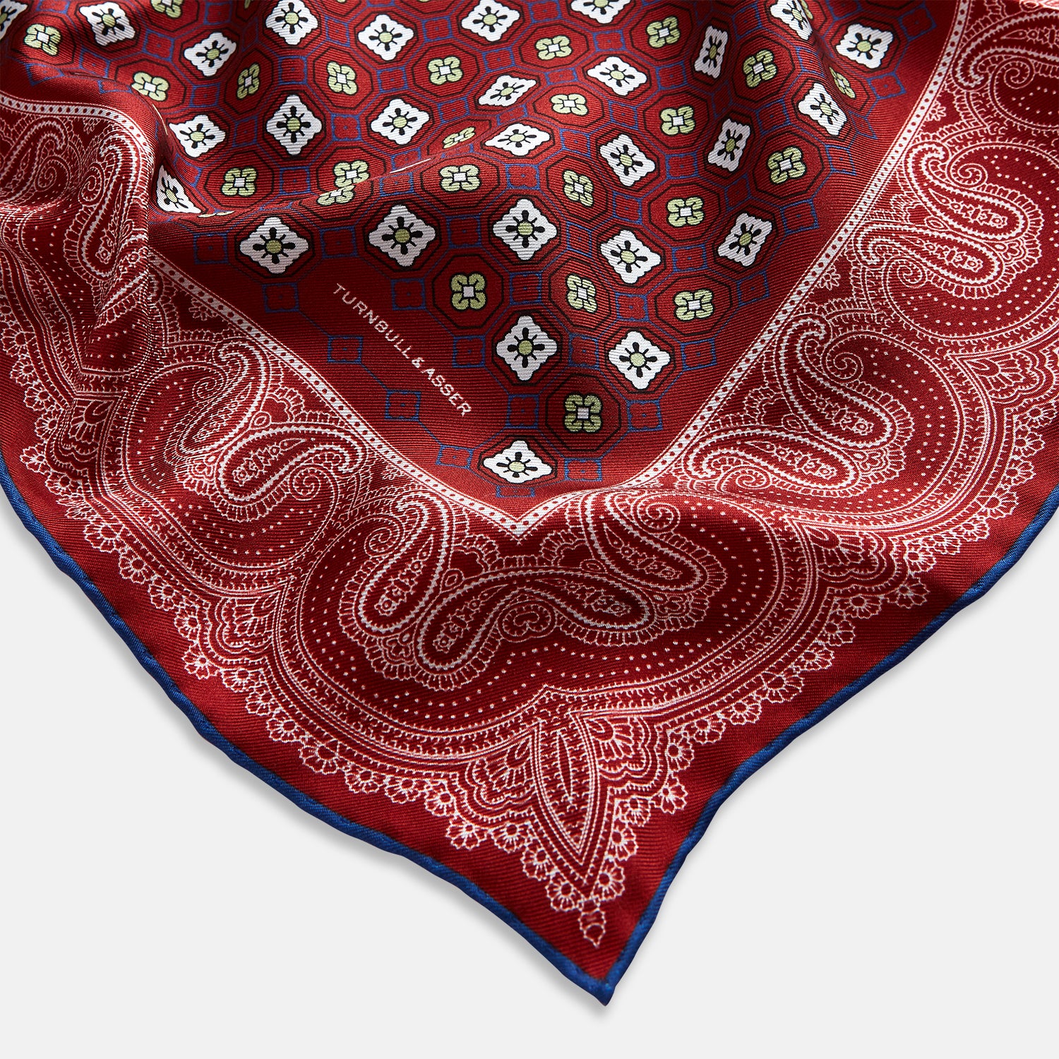 Image 2 of product The Turnbull & Asser Red Floral Tile and Paisley Silk Pocket Square features red silk with white and green paisley and geometric patterns, a blue border, a folded corner revealing both sides, branding in white, and is handcrafted in England.