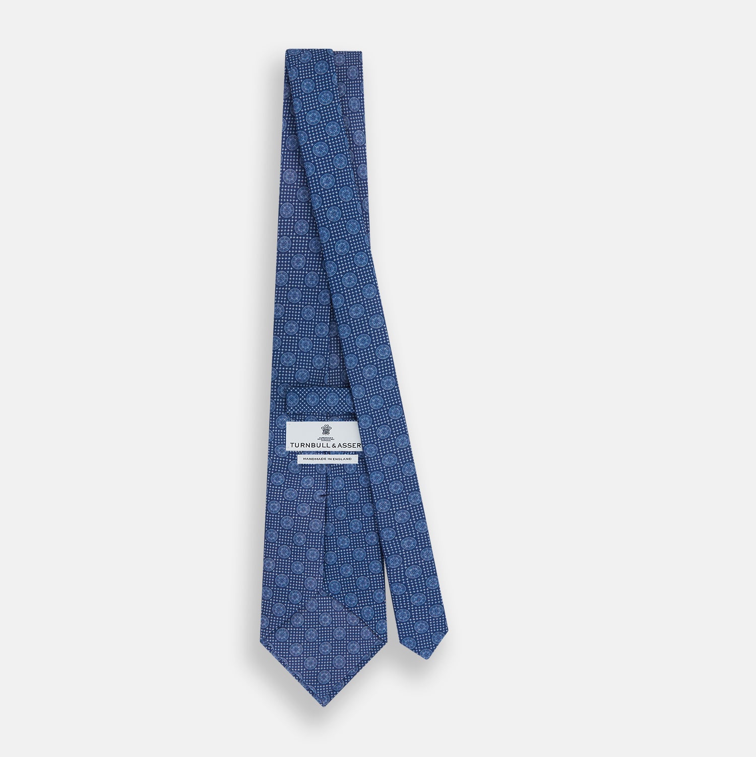 Image 4 of product The Turnbull & Asser Azzurro Medallion Silk Tie, rolled up and displayed on a white background, features small geometric medallion motifs in lighter blue tones.