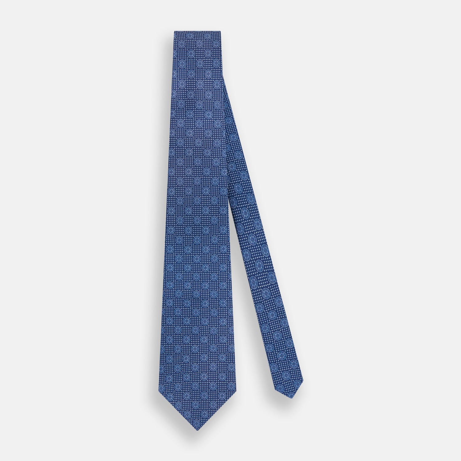 Image 2 of product The Turnbull & Asser Azzurro Medallion Silk Tie, rolled up and displayed on a white background, features small geometric medallion motifs in lighter blue tones.