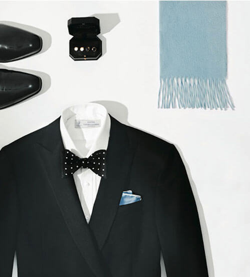 Flat lay of a black tuxedo with a white shirt, black polka dot bow tie, light blue pocket square, black dress shoes, a blue fringed scarf, and a black ring box with rings inside.