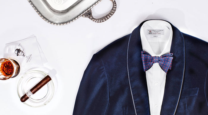 A navy blue tuxedo jacket with a white shirt and polka dot bowtie, next to a silver tray, a glass of whiskey, a cigar in an ashtray, and a white napkin on a white surface.