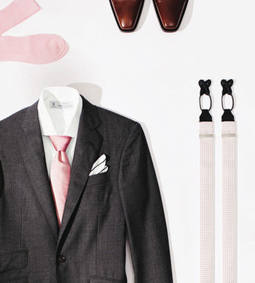 A dark gray suit with a white shirt, pink tie, and white pocket square is laid out alongside pink socks, brown dress shoes, and a pair of suspenders on a white background.