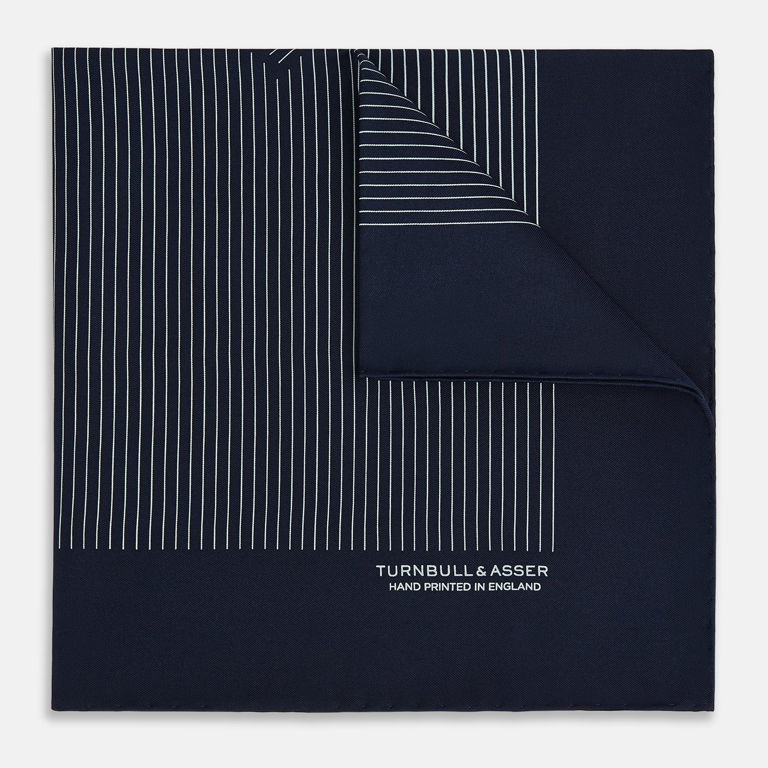 Image 1 of product The Navy and White Stripe Silk Pocket Square by Turnbull & Asser features thin white vertical stripes and "TURNBULL & ASSER HAND PRINTED IN ENGLAND" in one corner, with a folded edge showing the reverse of this elegant silk accessory.