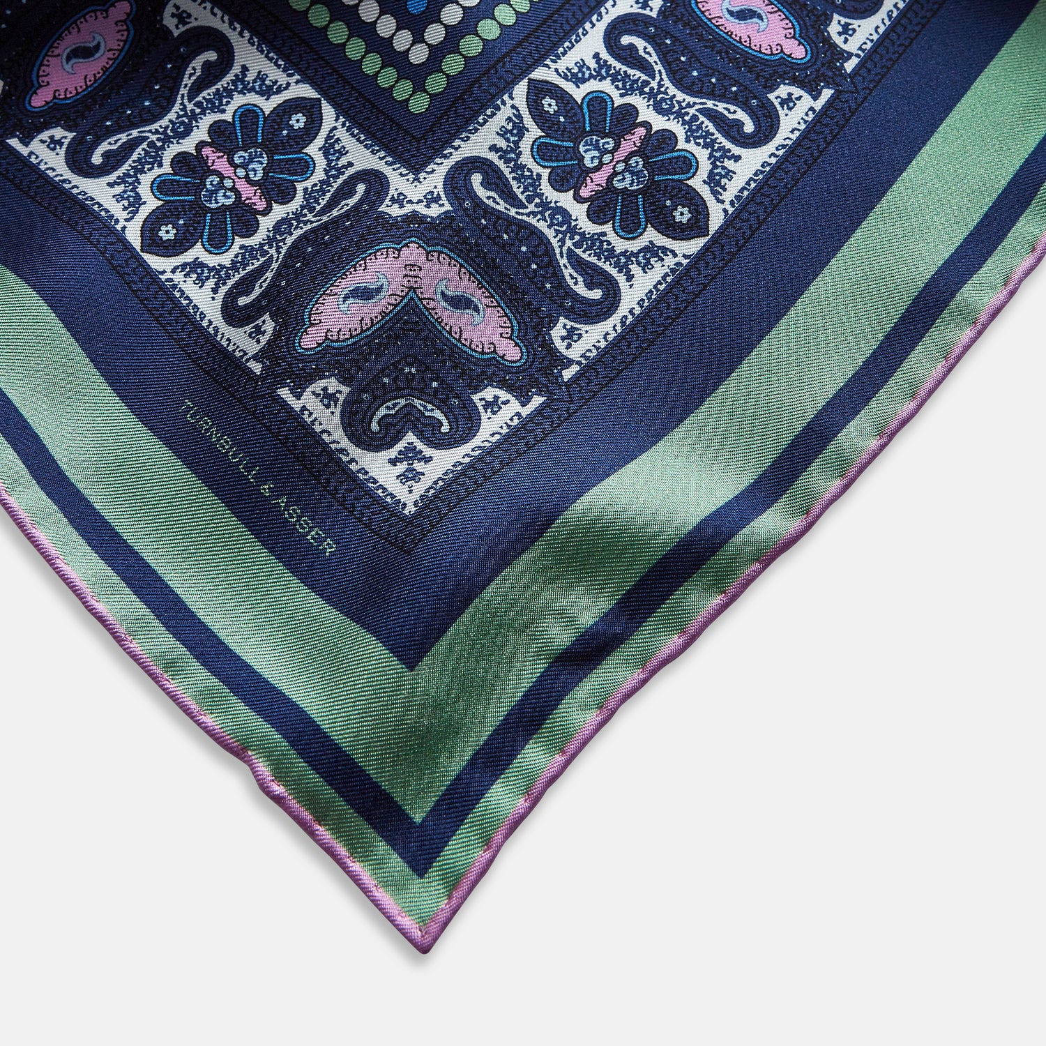 Image 2 of product The Turnbull & Asser Oxford Blue Dots and Paisley Silk Pocket Square is handcrafted in England and features a blue, green, and white polka dot pattern with a paisley and floral border, plus the brand’s signature logo in the corner.