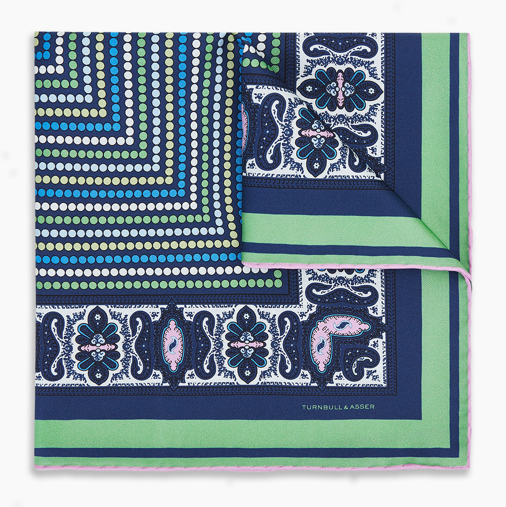 Image 1 of product The Turnbull & Asser Oxford Blue Dots and Paisley Silk Pocket Square is handcrafted in England and features a blue, green, and white polka dot pattern with a paisley and floral border, plus the brand’s signature logo in the corner.