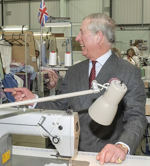A man in a gray suit smiles and points while standing next to an industrial sewing machine in a garment factory. The workspace has clothing racks, spools of thread, and a small British flag in the background.