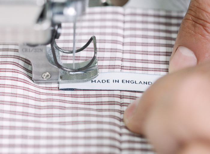 Close-up of a sewing machine attaching a MADE IN ENGLAND label to a piece of checkered fabric, with a persons hand guiding the fabric.