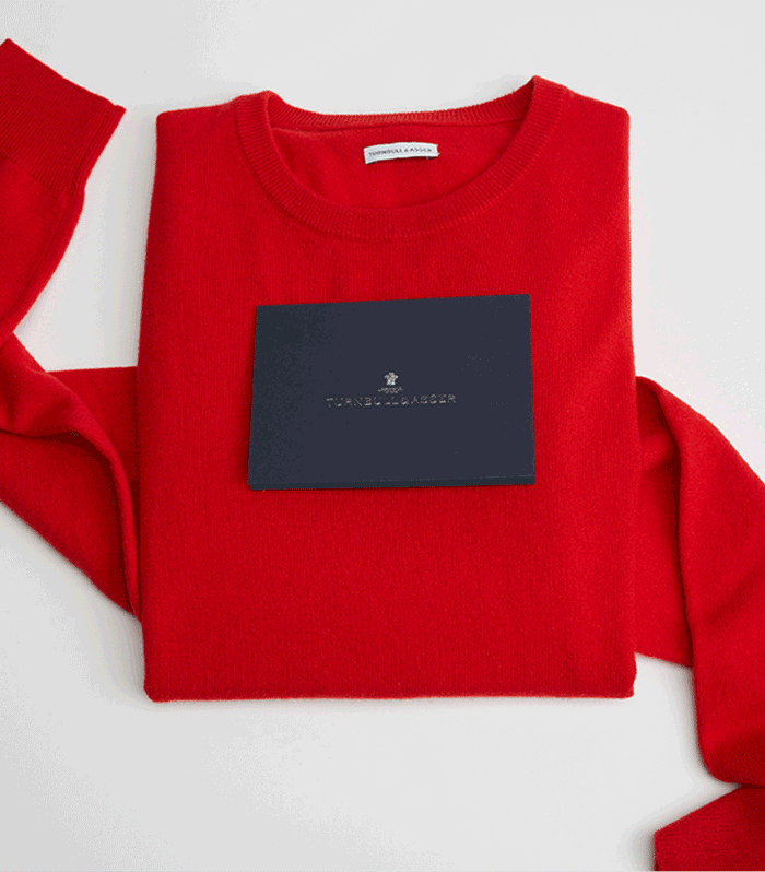 A bright red sweater neatly folded with a navy blue card from Turnbull & Asser placed on top, set against a white background.