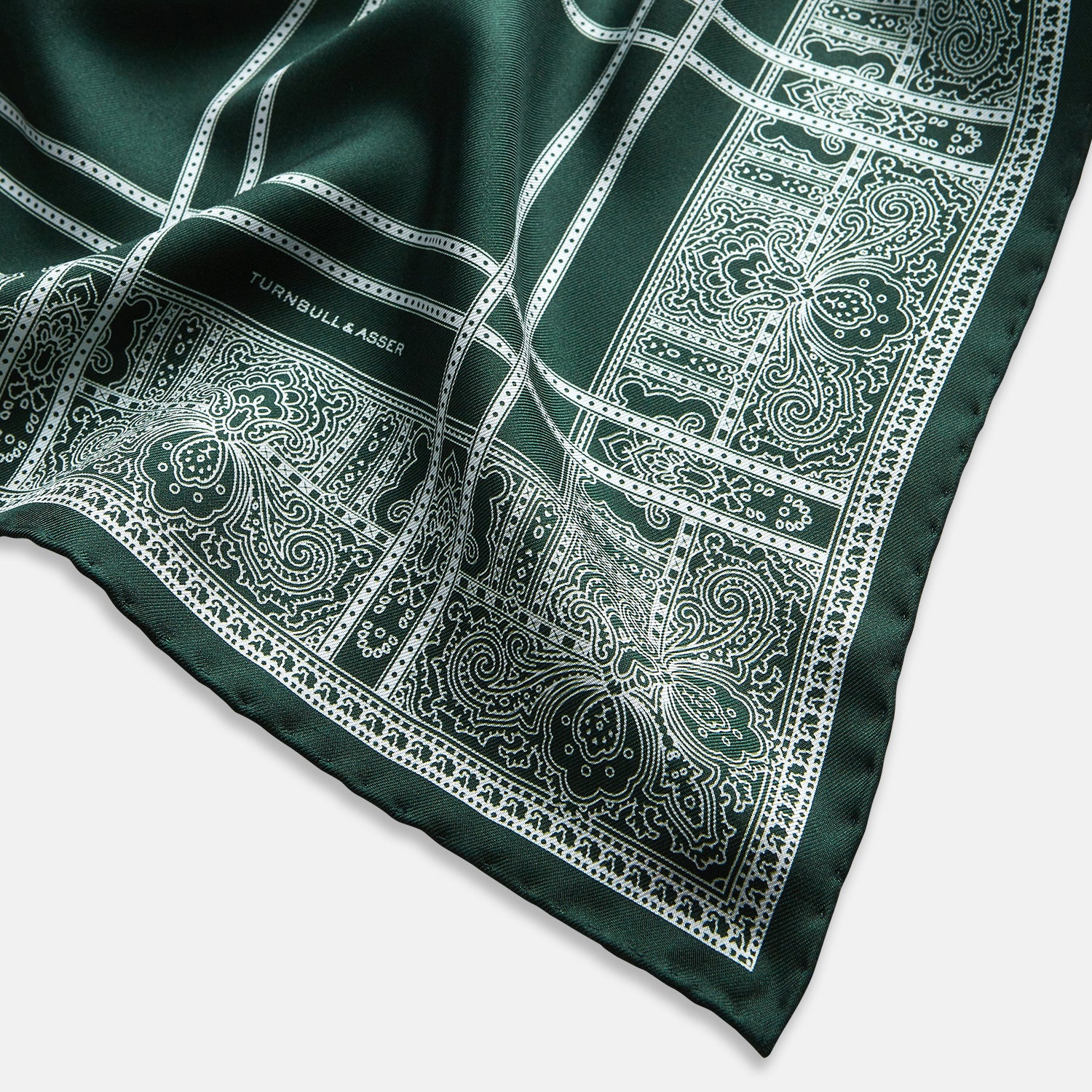 Image 2 of product Turnbull & Asser’s Forest Green Paisley Bandana Patterned Silk Pocket Square is handcrafted in England, featuring an intricate white geometric and paisley design with the brand name printed in one corner.