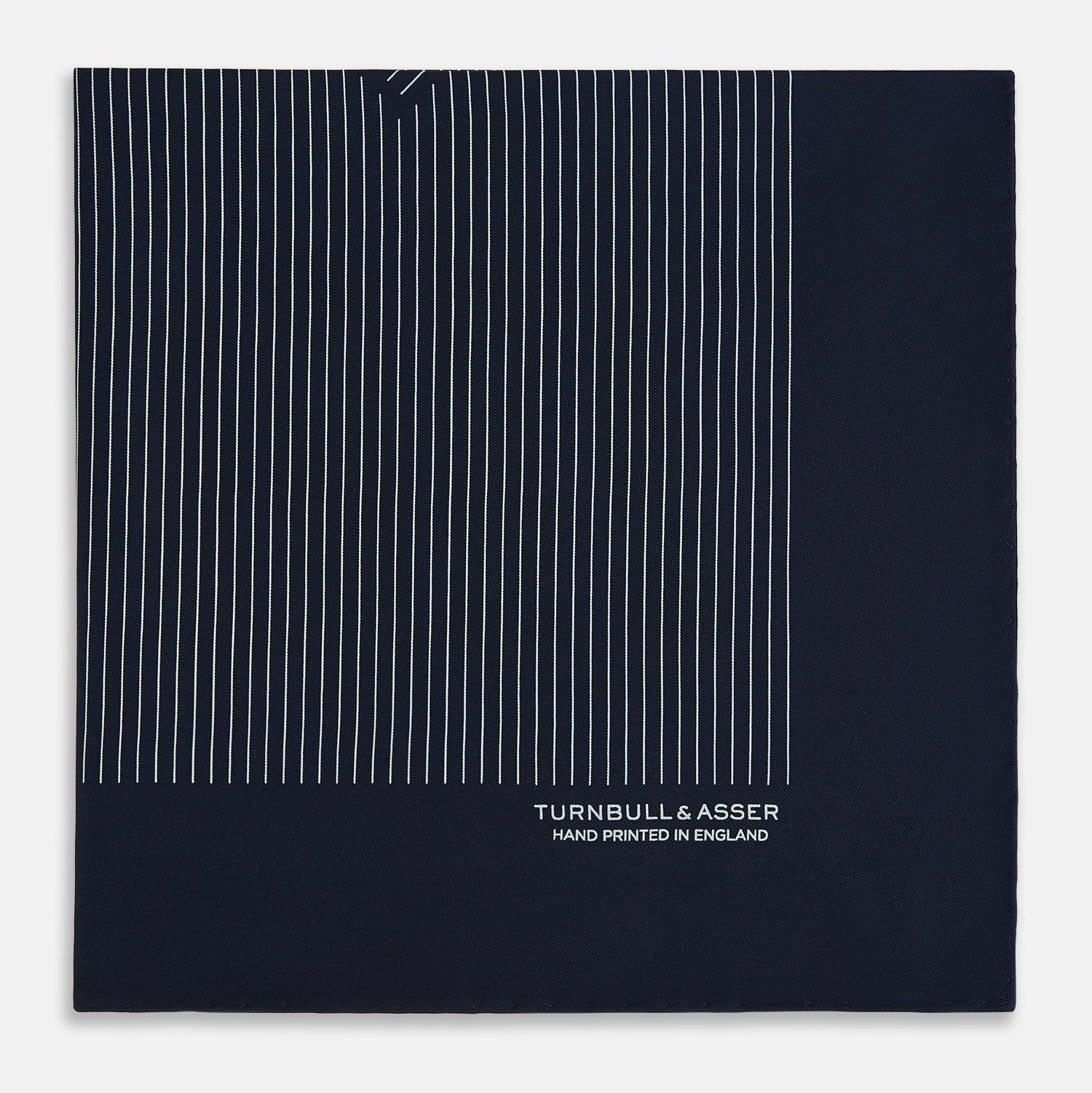 Image 4 of product The Navy and White Stripe Silk Pocket Square by Turnbull & Asser features thin white vertical stripes and "TURNBULL & ASSER HAND PRINTED IN ENGLAND" in one corner, with a folded edge showing the reverse of this elegant silk accessory.