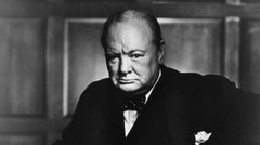 A black-and-white portrait of an older man in a suit and bow tie, looking serious, seated in front of a wooden panel background.