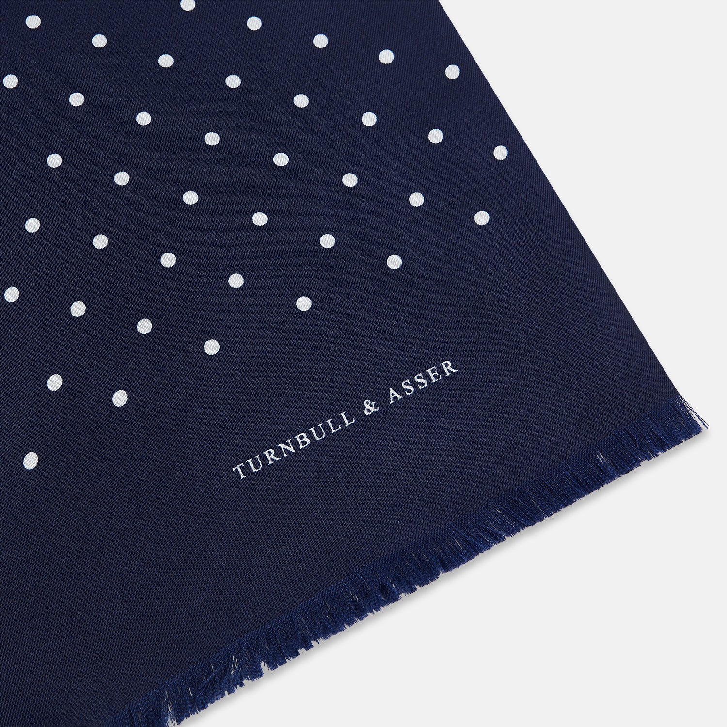 Image 2 of product The Navy and White Spotted Silk Scarf by Turnbull & Asser features white polka dots, a fringed edge, and the brand's name printed near the bottom on both sides. It is neatly folded at the top.