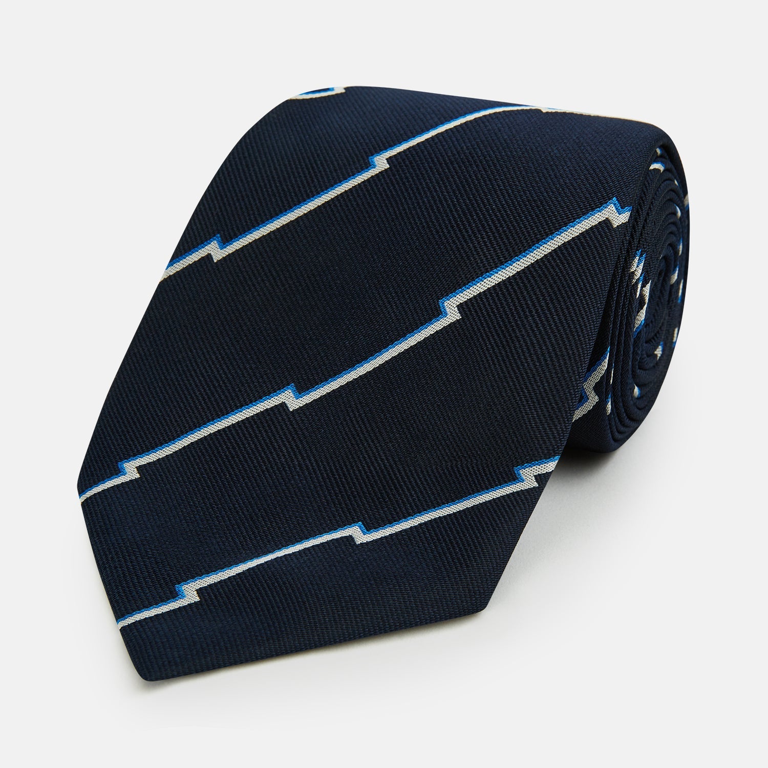 Image 1 of product The Turnbull & Asser Navy Broken Shadow Stripe Silk Tie features British excellence with a light blue and white zigzag pattern and is shown rolled up on a plain white background.