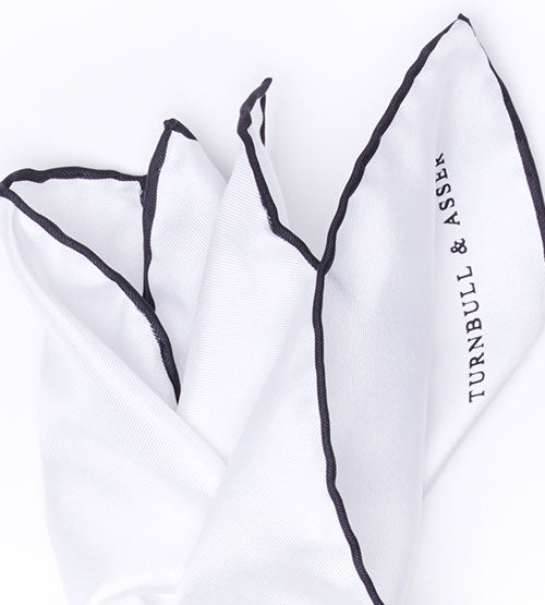 A white pocket square with black edging, partially folded, featuring the text TURNBULL & ASSER printed along one edge.