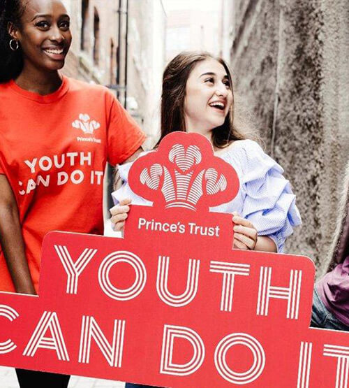 Two young women smiling and holding red signs that read “Prince’s Trust” and “Youth Can Do It” in a narrow outdoor alley. One woman wears a red t-shirt with the same slogan.