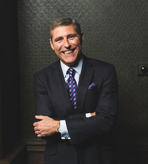 A man in a dark suit and patterned tie stands smiling with arms crossed in front of a textured dark wall. He has short hair and is wearing a watch and pocket square.
