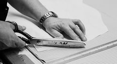 A person uses large metal scissors to cut fabric along a marked pattern on a worktable, with one hand holding the fabric steady and wearing a watch.