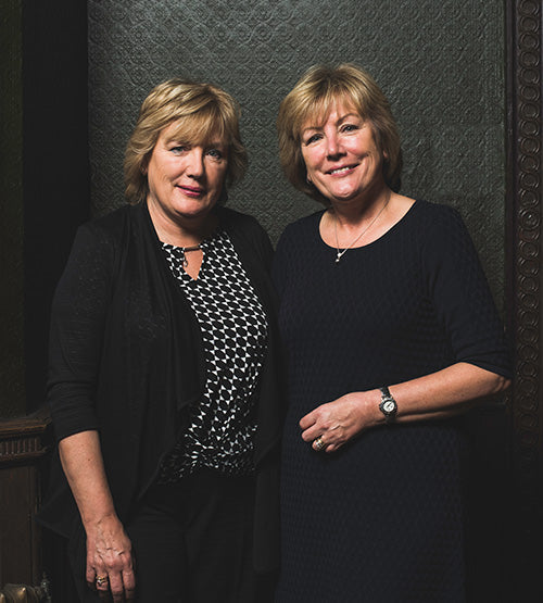 Two women with short blonde hair stand side by side, smiling for the camera. They are dressed in dark clothing and are positioned in front of a textured dark background.