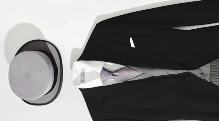 A formal black suit with a white dress shirt, grey vest, and patterned tie is laid out next to a grey bowler hat on a white surface.