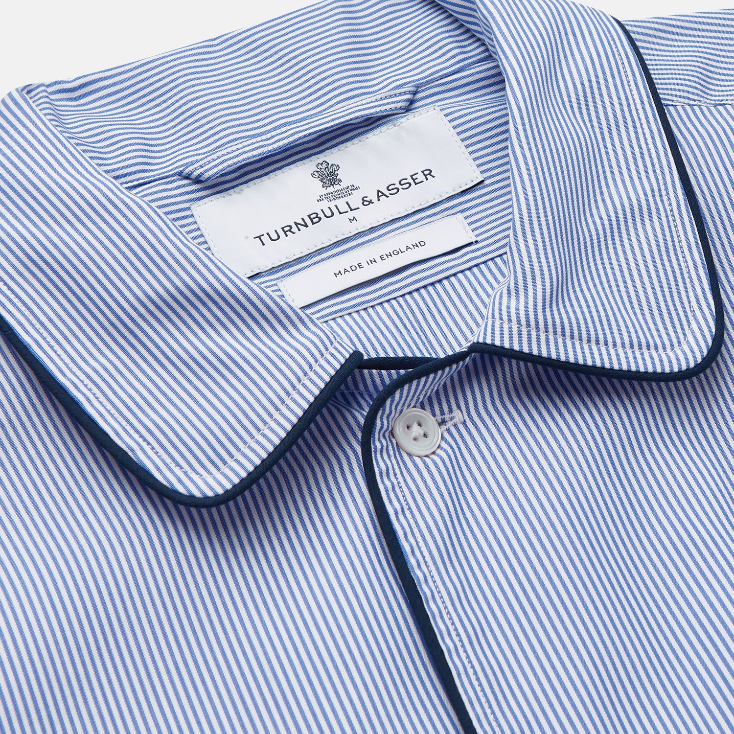 Image 2 of product The Turnbull & Asser Blue Fine Bengal Stripe Piped Cotton Nightshirt features long sleeves, a chest pocket, black collar piping, curved hem, and side slits. It is displayed laid flat on a white background.