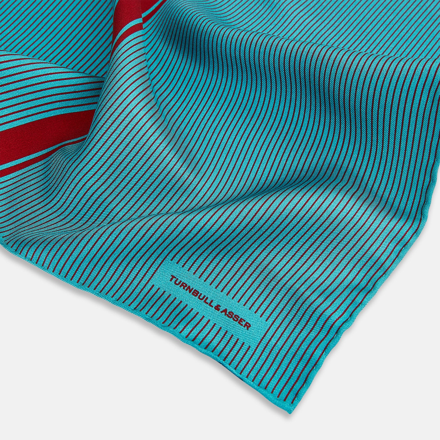 Image 2 of product The Turnbull & Asser Turquoise and Red Stripe Silk Pocket Square features thin navy stripes with a bold vertical red stripe, partially folded to reveal the reverse pattern and the brand's logo, showcasing its British heritage.