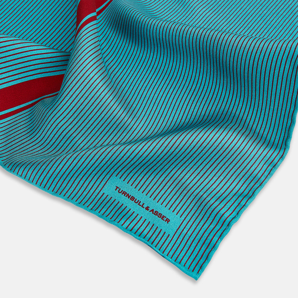 The Turnbull & Asser Turquoise and Red Stripe Silk Pocket Square features thin navy stripes with a bold vertical red stripe, partially folded to reveal the reverse pattern and the brand's logo, showcasing its British heritage.