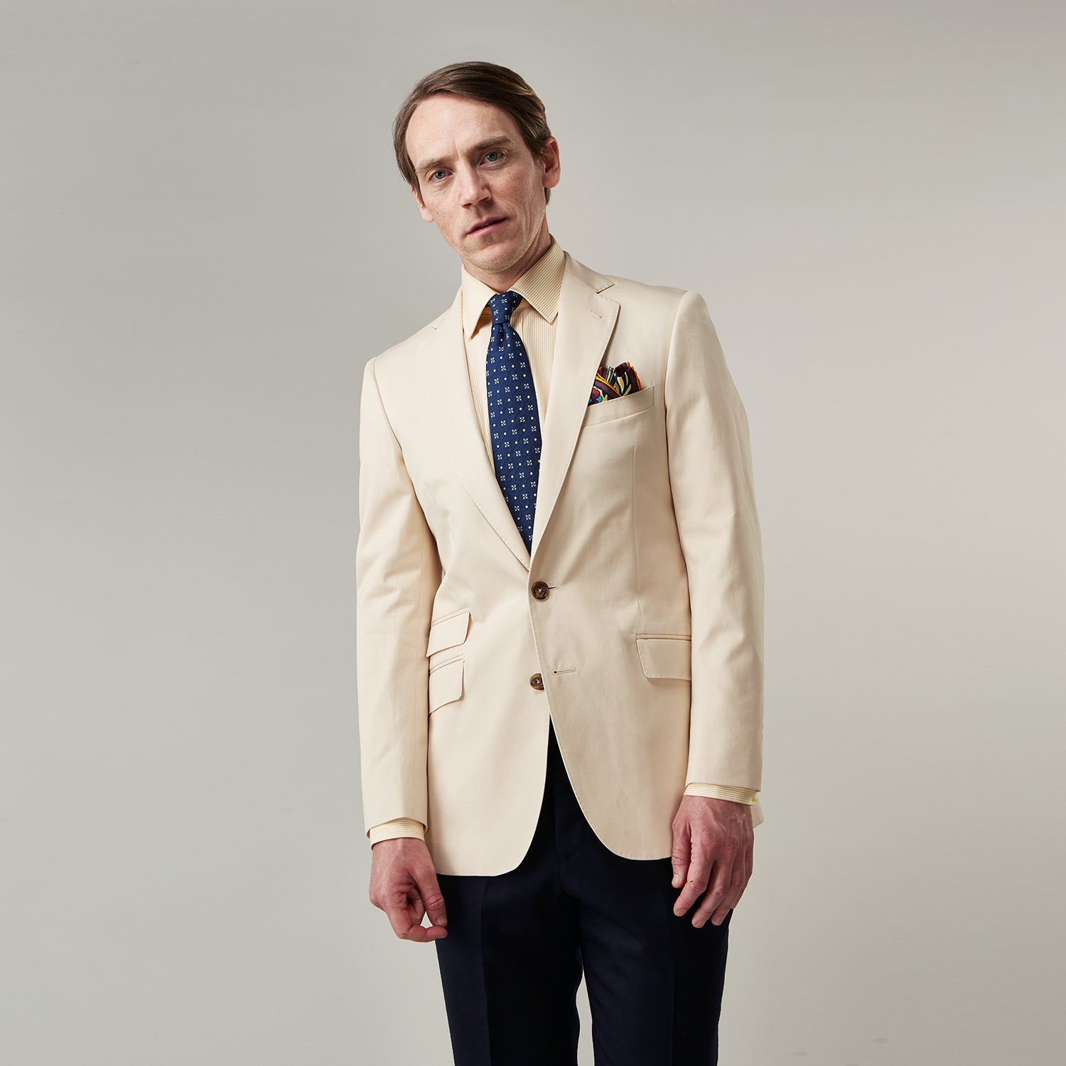 View 1 of product A man in a Turnbull & Asser Beige Cotton Barrington Blazer, light yellow shirt, blue patterned tie, and dark trousers stands against a plain light background, with a pocket square visible and a neutral expression.