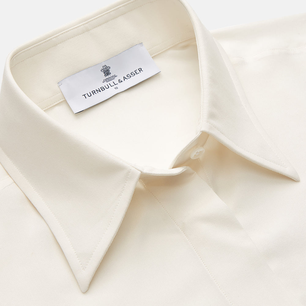 The Cream Silk Tailored Bianca Shirt by Turnbull & Asser features long sleeves, a pointed collar, and curved hem in soft-touch peace silk with a tailored fit. Shown laid flat on a white background.