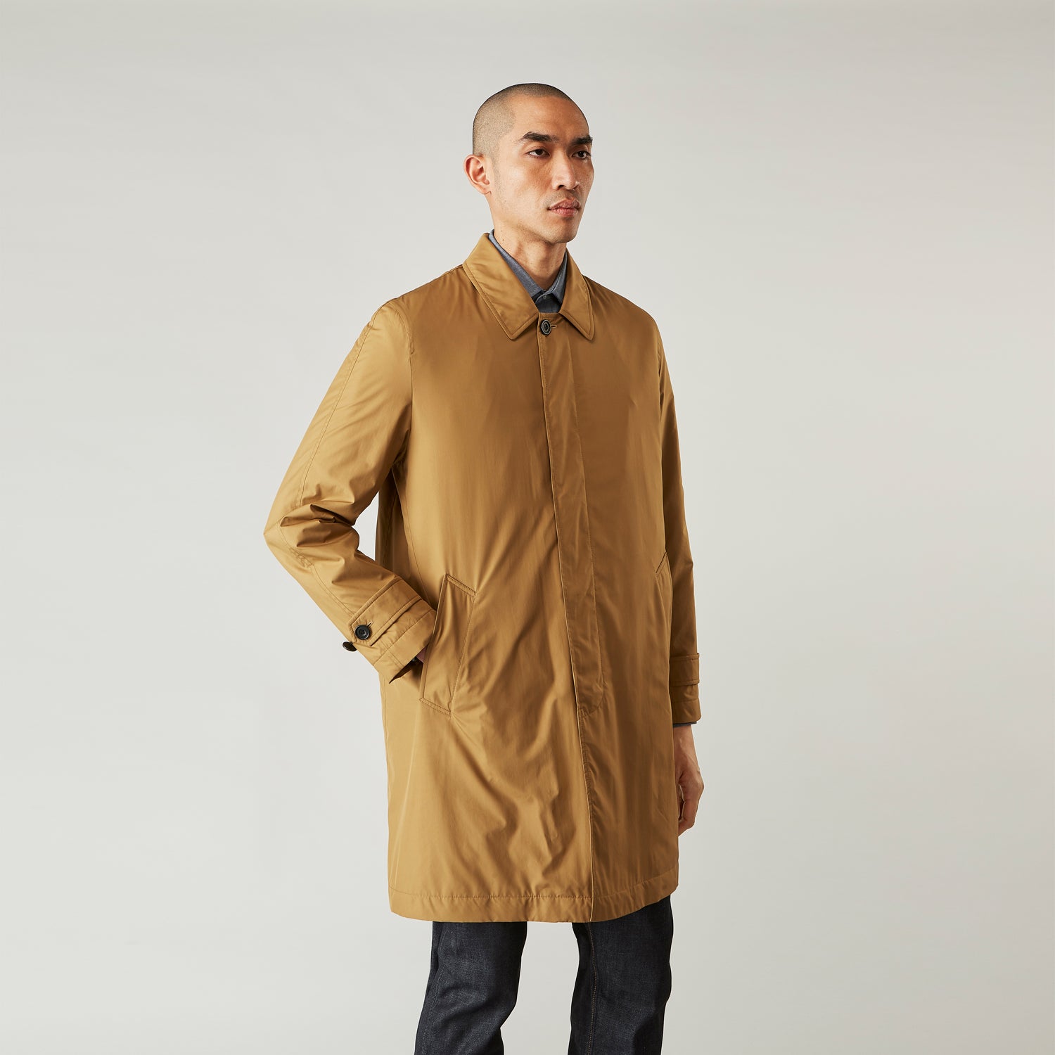View 1 of product A person wears the Turnbull & Asser Camel Quintern Sealup Raincoat with Detachable Liner, paired with dark pants, standing against a plain light gray background, showing the sleek technical rainwear style with one hand in a pocket.