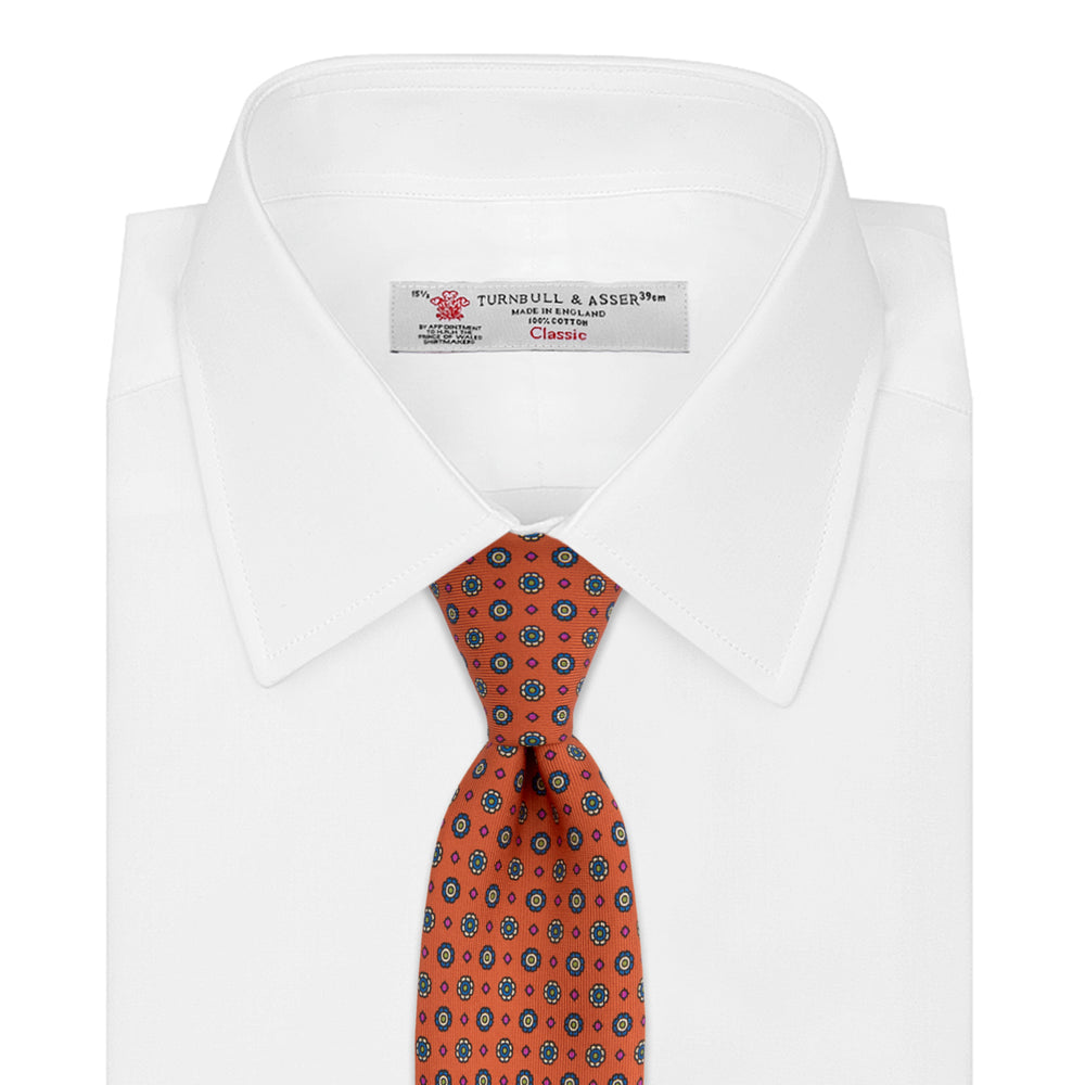 The Great Gatsby Orange Printed Silk Tie by Turnbull & Asser, from the Legends collection, features a repeating pattern of small blue, white, and yellow floral motifs—an ideal summer accessory—shown rolled on a white background.