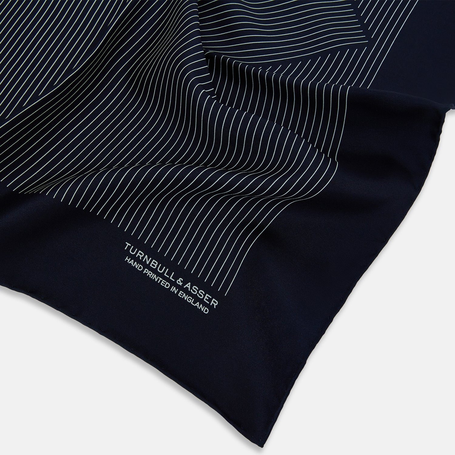 View 2 of product The Navy and White Stripe Silk Pocket Square by Turnbull & Asser features thin white vertical stripes and "TURNBULL & ASSER HAND PRINTED IN ENGLAND" in one corner, with a folded edge showing the reverse of this elegant silk accessory.