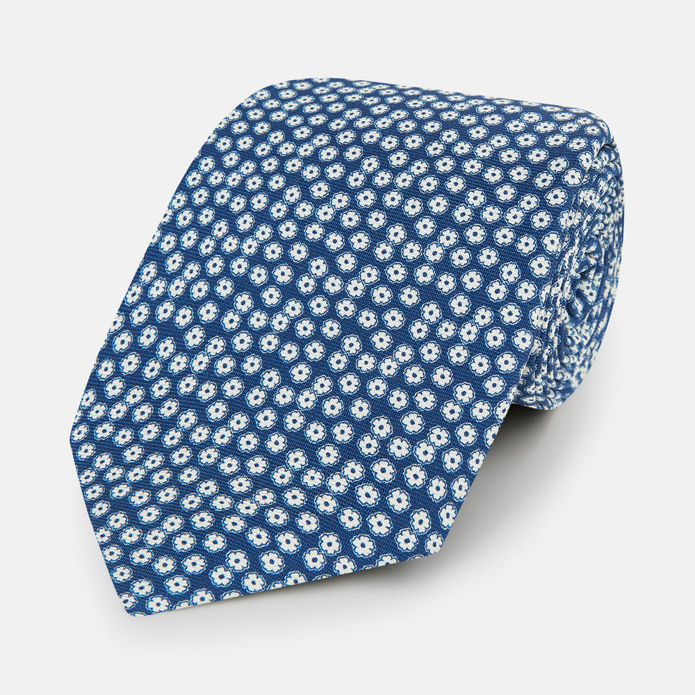 The Turnbull & Asser Azzurro Flower Patterned Silk Tie features a repeating motif of small white flowers with dark centers on blue silk, rolled up and displayed against a plain white background.