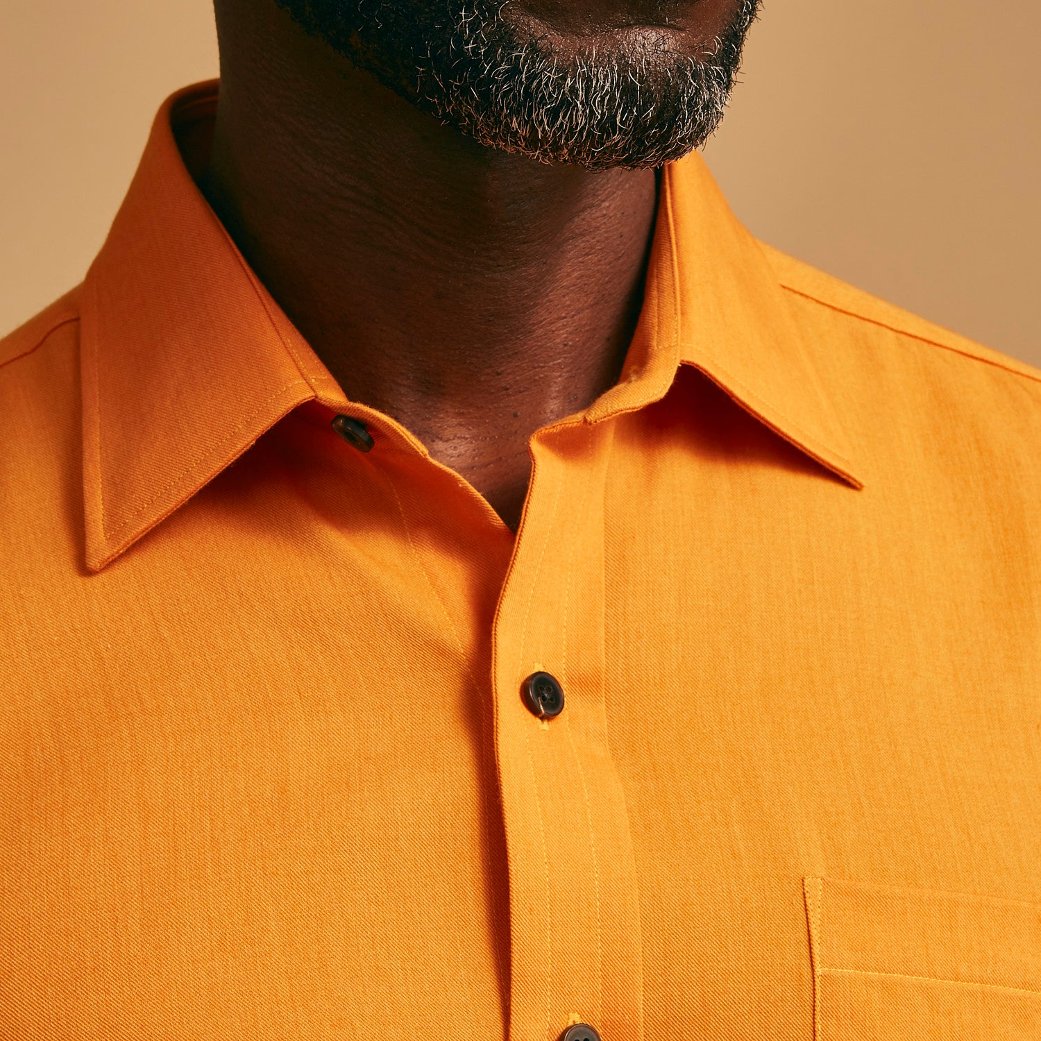View 4 of product A man stands against a tan background, wearing a Turnbull & Asser Orange Weekend Fit Cotton & Wool Shirt with Derby Collar and 1 Button Cuffs, tucked into dark blue dress pants with a black belt and shoes. He has one hand in his pocket.