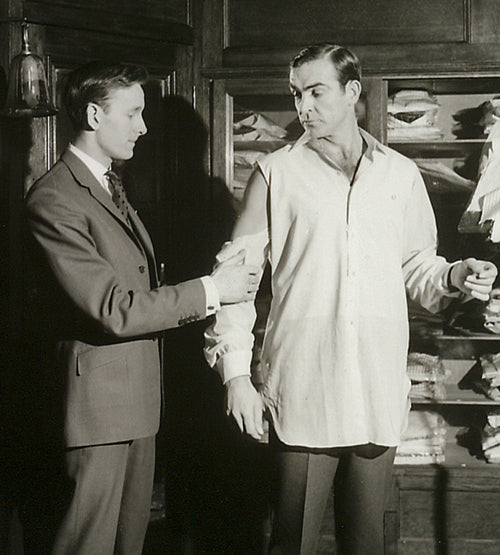A tailor in a suit measures the sleeve of a man wearing a shirt in front of shelves stacked with neatly folded shirts. The man being measured looks down at his sleeve while the tailor adjusts it.