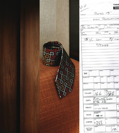A mannequin with a patterned tie is next to a tailored suit order form labeled “Bond 19” with measurements, fabric details, and production notes visible.