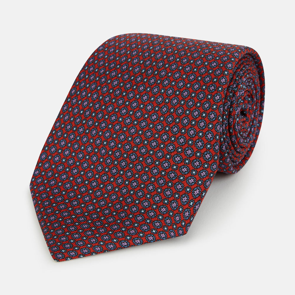 The Red Multi-Floral Silk Tie by Turnbull & Asser features a blue and white geometric pattern and is shown rolled up against a plain white background.