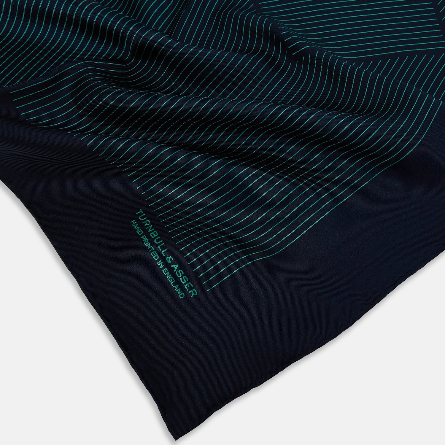 View 2 of product A Navy and Petrol Green Stripe Silk Pocket Square by Turnbull & Asser, featuring thin petrol green stripes and "Turnbull & Asser Hand Printed in England" in the bottom right corner. One corner is folded to display hand-rolled edges.