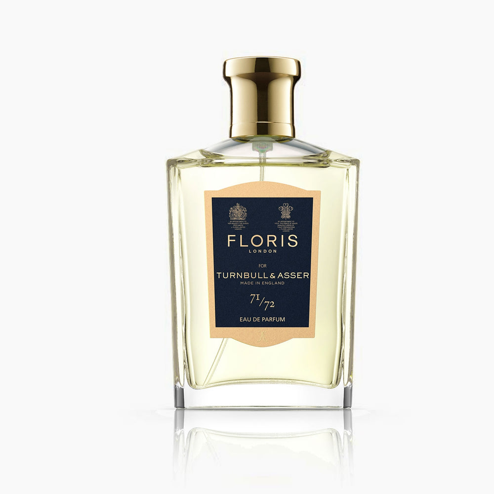 A bottle of Floris 71/72 Eau De Parfum by Turnbull & Asser, a woody aromatic scent, stands beside its navy blue and gold box. The 100 ml bottle features a gold cap in this exclusive collaboration.
