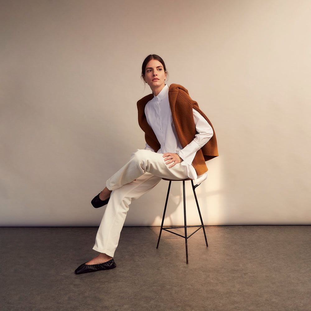 A person with curly hair wears the Turnbull and The Deck White Cotton Minimalist Shirt by Turnbull & Asser, paired with pants. Posed against a plain background, their minimalist silhouette is highlighted in this black and white image.