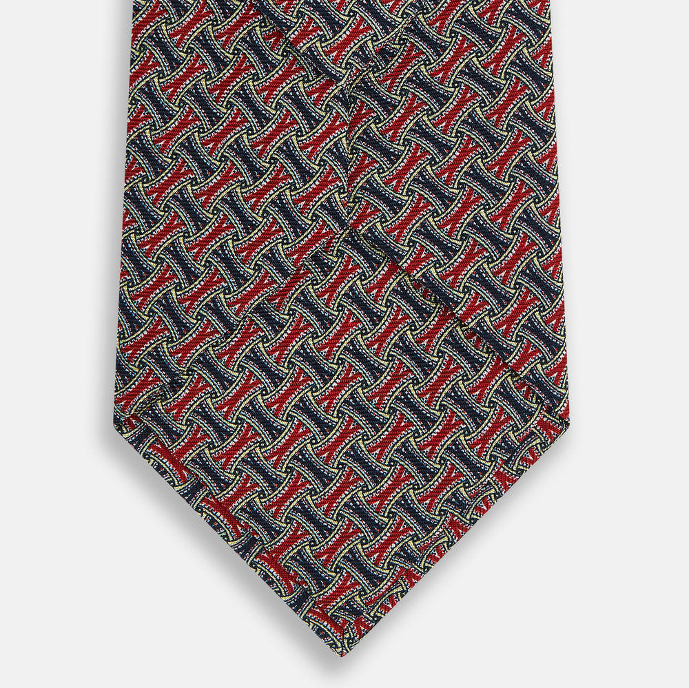 The Turnbull & Asser Red and Navy Weave-Patterned Printed Silk Tie features an intricate geometric design in red, blue, and white. Displayed rolled-up against a plain white background, this classic tie is crafted from luxurious silk.