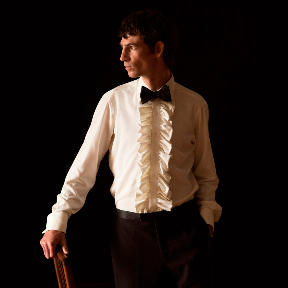 A man in a Cream Silk Regular Fit Ruffle-Front Wilfred Shirt by Turnbull & Asser stands against a dark background, one hand on a chair, gazing thoughtfully to the side.