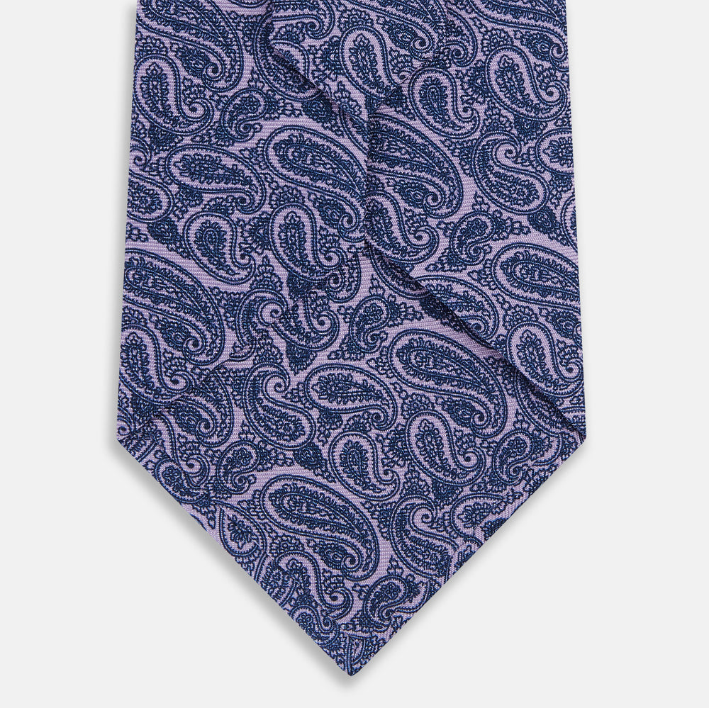 The Turnbull & Asser Lilac Paisley Silk Tie is shown rolled up, featuring a blue paisley pattern on a light purple background and displayed on a white surface.