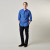 View 1 of product A man faces forward with a neutral expression, hands in his pockets, wearing the Turnbull & Asser Plain Indigo Linen Weekend Fit Shirt with a stand collar, styled with dark pants and black shoes against a plain light background.