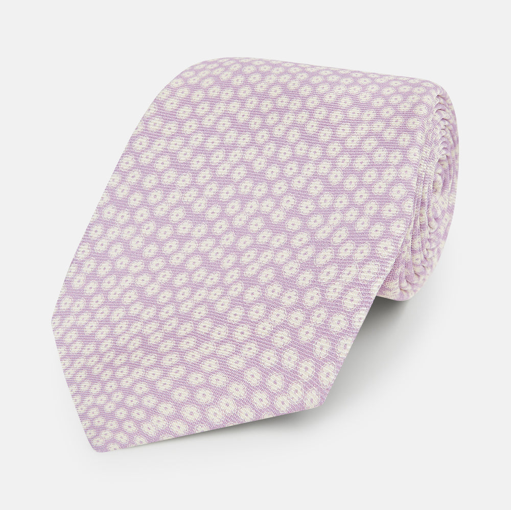 The Turnbull & Asser Lilac Flower-Patterned Printed Silk Tie, featuring a light lilac hue and small white circular designs, is shown rolled up against a plain white background.