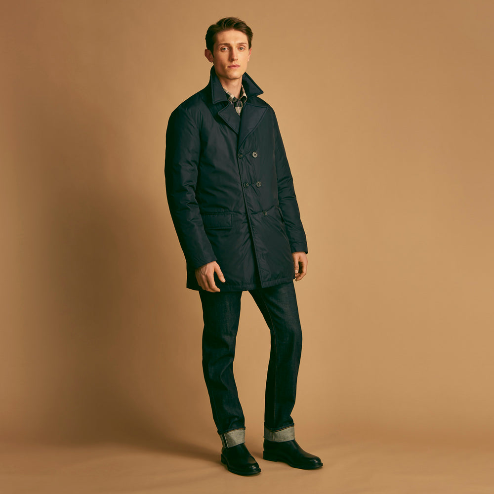 A man stands against a beige background, wearing the Turnbull & Asser Navy Burrows Double Breasted Coat over a plaid shirt and mauve cardigan, paired with dark rolled-up jeans and black shoes, looking directly at the camera with a neutral expression.