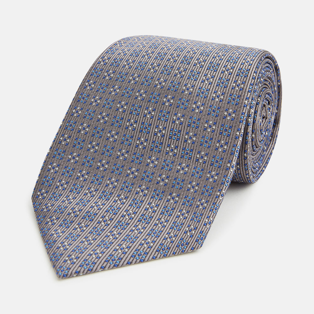 The Turnbull & Asser Silver and Pale Blue Multi Dot Silk Tie features elegant small dots on light brown silk, ideal for modern office wear, shown rolled up against a white background.