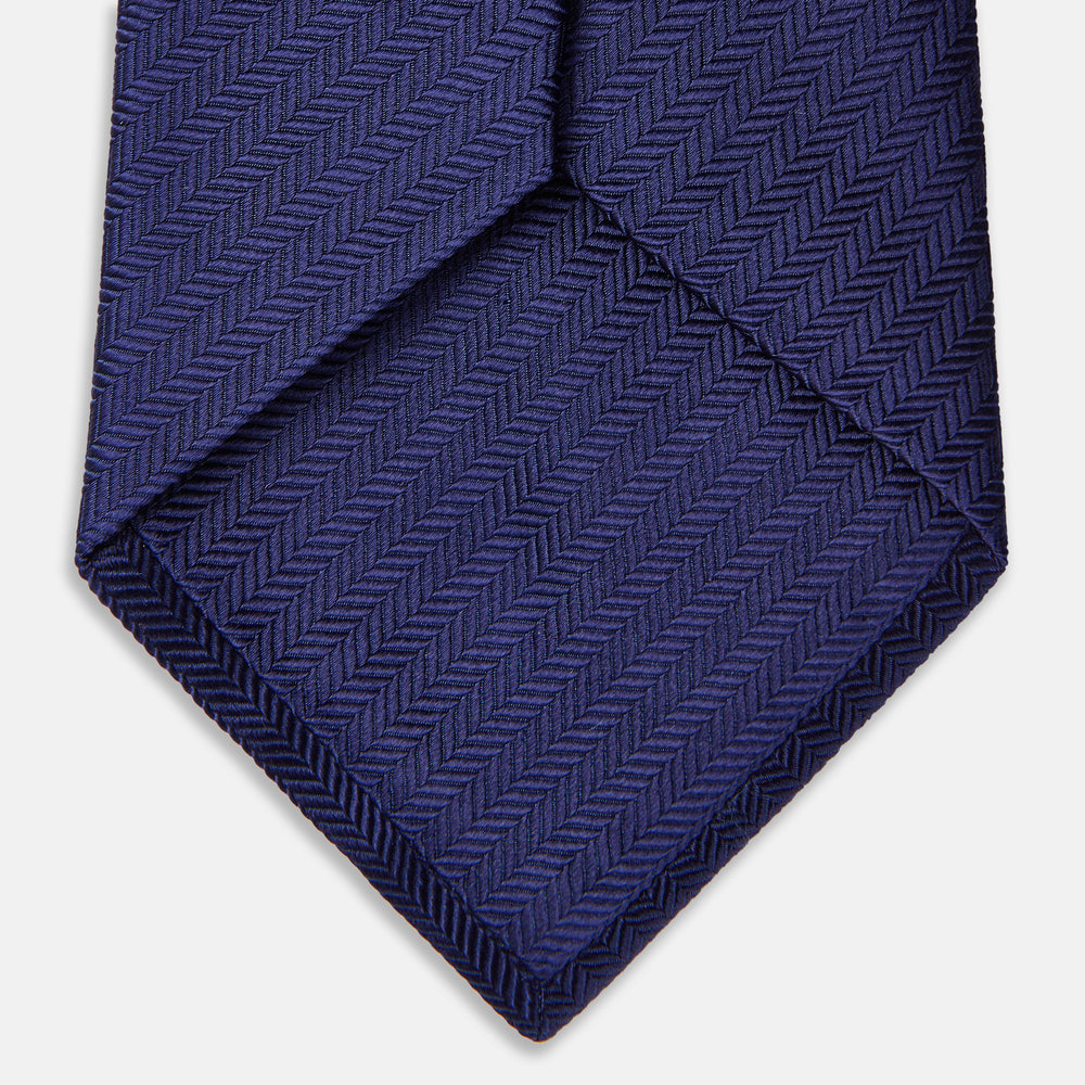 The PLAIN HERRINGBONE NAVY TIE by Turnbull & Asser, crafted from silk with a subtle pattern, is elegantly rolled and displayed against a plain white background.