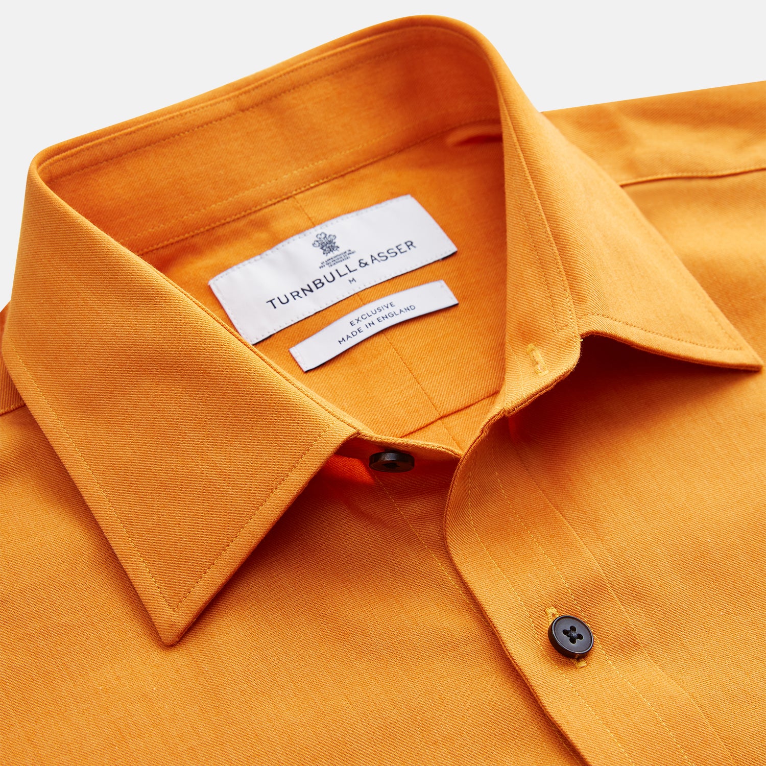 View 3 of product A man stands against a tan background, wearing a Turnbull & Asser Orange Weekend Fit Cotton & Wool Shirt with Derby Collar and 1 Button Cuffs, tucked into dark blue dress pants with a black belt and shoes. He has one hand in his pocket.