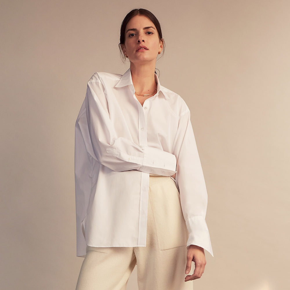 A woman stands barefoot indoors against a neutral wall, wearing Turnbull & Asser Women's Cream Cashmere Knitted Tabitha Bottoms and a light brown sweater. Soft sunlight casts a gentle shadow on the wall beside her.