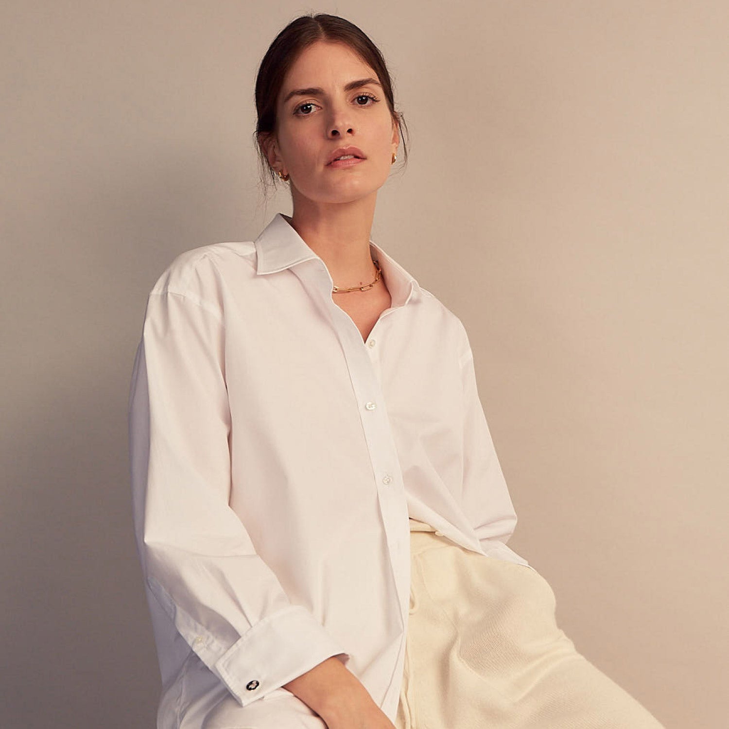 View 6 of product A woman with straight brown hair pulled back wears the Turnbull & Asser White Cotton Marlene Shirt, featuring classic menswear details, paired with high-waisted cream trousers against a neutral background.